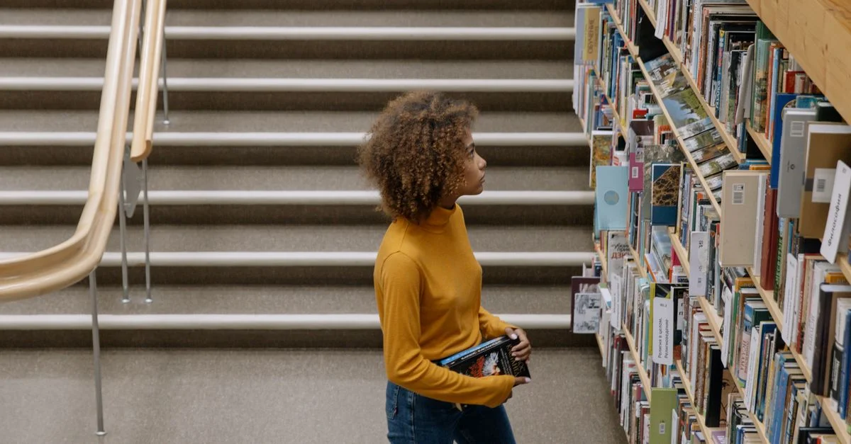 Jeune femme en pull jaune feuilletant des livres dans une bibliothèque avec des étagères et des escaliers.